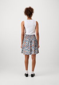 Donna con capelli ricci, che indossa un top senza maniche bianco e una gonna floreale al ginocchio, in piedi di spalle su uno sfondo semplice.