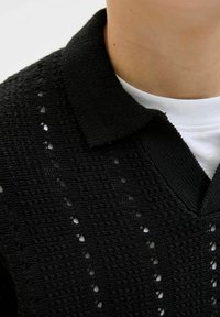 Polo en tricot noir avec un motif texturé et de petits détails en œillets. Le col est uni avec un design en V et un look superposé.