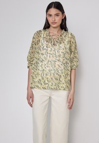 Gelbe Bluse mit floralem Muster, Schnürsaum, Puffärmeln und einem ganzflächigen botanischen Druck, kombiniert mit cremefarbener, weit geschnittener Hose.
