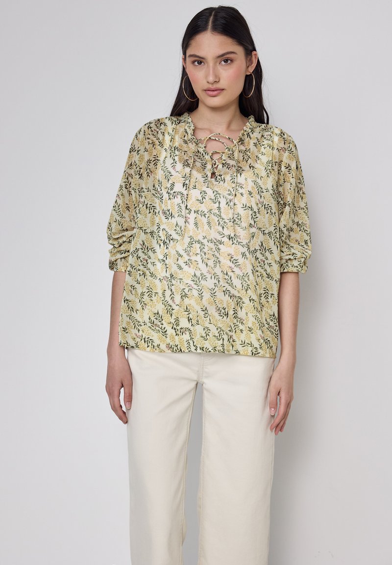 Gelbe Bluse mit floralem Muster, Schnürsaum, Puffärmeln und einem ganzflächigen botanischen Druck, kombiniert mit cremefarbener, weit geschnittener Hose.