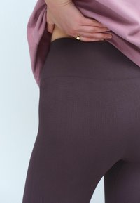 Ljubičaste rebraste tajice s visokim pojasom, u kombinaciji s svijetloružičastim gornjim dijelom, djelomično prekrivaju tajice na neutralnoj pozadini.