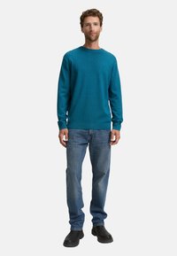 Türkiser Pullover mit langen Ärmeln und rundem Ausschnitt, kombiniert mit hellblauen Jeans und schwarzen Stiefeletten. Glatte Textur, lässige Passform.