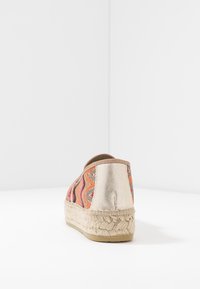 Espadrille-sko med vävd beige plattformssula, flerfärgad zigzag-tyg och metallisk gyllene hälflik, visad bakifrån mot vit bakgrund.