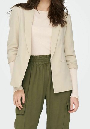 Kobieta w beżowej marynarce na jasnobeżowym swetrze i oliwkowozielonych, wysokich spodniach typu cargo, stojąca z rękami swobodnie opuszczonymi wzdłuż ciała.