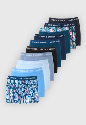 Stojący mężczyzna w niebieskich bokserkach z tropikalnym nadrukiem i paskiem '"Jack & Jones"', patrzy w bok na tle białej ściany.
