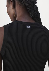 Haut noir sans manches à côtes porté, avec une petite étiquette « Moschino Jeans » au haut du dos et des cheveux tressés tirés en arrière.