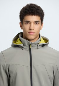 Veste à capuche gris clair avec une doublure intérieure jaune, fermeture éclair à l'avant et capuche avec cordon de serrage ajustable. Texture lisse et design minimaliste.