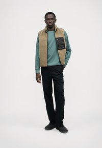 Γκρι φλις γιλέκο πάνω από τυρκουάζ μακρυμάνικο πουκάμισο, μαύρο παντελόνι και μαύρα παπούτσια. Το γιλέκο έχει μια μπροστινή τσέπη με μια μικρή ετικέτα. Απλώς φόντο.