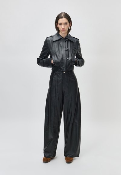 Γυναίκα που φοράει μαύρο cropped δερμάτινο μπουφάν και δερμάτινο παντελόνι με φαρδιά ψηλή μέση, μαζί με καφέ παπούτσια, στέκεται με τα χέρια στις τσέπες.