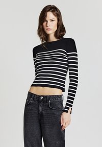 Giovane donna che indossa un maglione cropped a maniche lunghe a righe bianche e nere e jeans neri sbiaditi a vita alta, davanti a uno sfondo neutro.