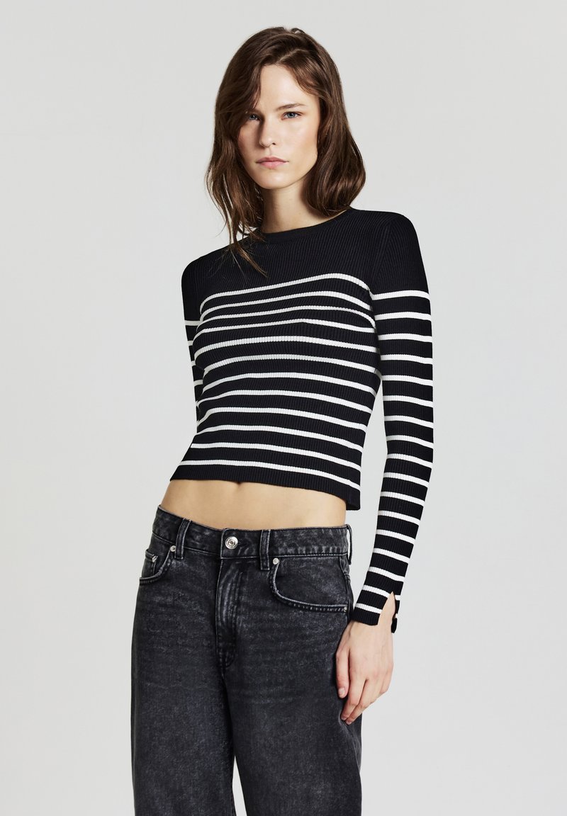 Giovane donna che indossa un maglione cropped a maniche lunghe a righe bianche e nere e jeans neri sbiaditi a vita alta, davanti a uno sfondo neutro.