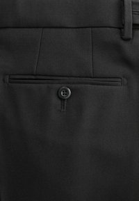 Black trousers featuring a back pocket with a horizontal seam and a single black button closure. Fabric has a smooth, textured finish.