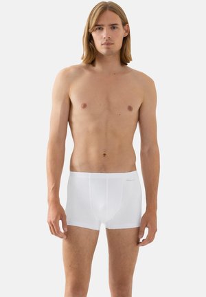 Junger Mann mit schulterlangem Haar, der steht und eine schlichte weiße Boxershorts vor einem hellgrauen Hintergrund trägt.