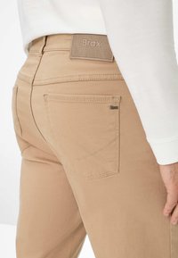 Beige Hose mit glatter Textur, ausgestattet mit einem gewebten Brax-Label, Gesäßtaschen und kontrastierenden Nähten.