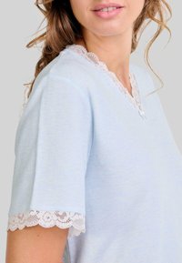Mujer vistiendo una blusa de manga corta azul claro con borde de encaje blanco en el escote y en los bordes de las mangas, contra un fondo sencillo.