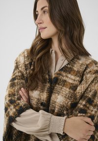 Texturerad brun och beige poncho med rutmönster, svarta accenter och knappknäppning. Bärs över en krämfärgad blus.