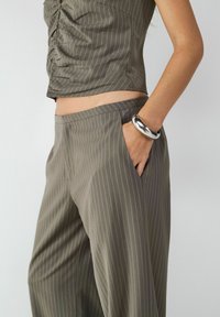 Szare spodnie w prążki o luźnym kroju z bocznymi kieszeniami, zestawione z dopasowaną bluzką; gładki materiał i prosty design. Srebrna bransoleta na dłoni.