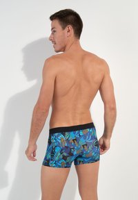 Herrenschwimmshorts mit einem blau-grünen tropischen Blattmuster auf dunklem Hintergrund. Elastischer schwarzer Bund, kurzes und enges Design.