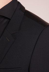 Blazer nero con tessuto testurizzato, accenti in pelle liscia sulle spalle, forma sartoriale e un sottile dettaglio metallico sul bavero.
