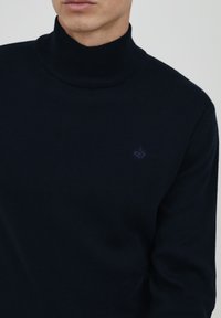Pulover turtleneck navy din țesătură tricotată în dungi. Dispune de un logo mic și discret pe piept. Textură netedă cu mâneci lungi.