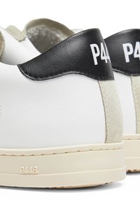 Gros plan de baskets blanches avec languettes de talon en cuir noir et détails en daim beige, avec marquage P448 sur le talon et la semelle.