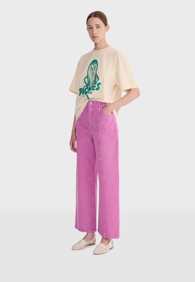 Femme aux cheveux attachés portant un t-shirt crème avec un motif vert et un pantalon large rose oversized, debout avec une main dans la poche.