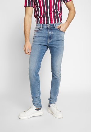 Homme portant un jean skinny bleu clair, des baskets blanches et une chemise à manches courtes à rayures verticales rouges, noires et blanches, main dans la poche.