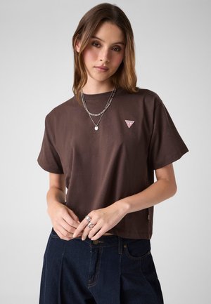 Giovane donna che indossa una maglietta marrone a maniche corte con un piccolo logo rosso, collane d'argento stratificate, anelli e jeans blu scuro su uno sfondo semplice.