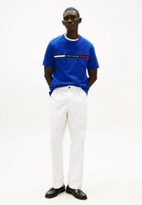 Blå bomulds T-shirt med hvide og røde vandrette striber, med teksten "TOMMY HILFIGER". Kombineret med hvide bukser og sorte sko.