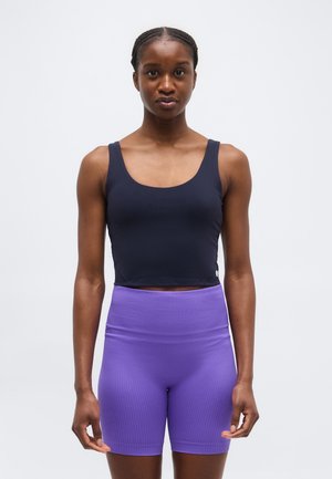 Žena stojaca v tmavomodrom krátkom tielku a vysokým pásom fialových rebrovaných cyklistických šortkách na jednoduchom pozadí.
