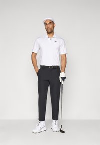 Mand i hvid golfkasket, hvid polo, sorte bukser og hvide golfsko holder en putter med den højre hånd iført en handske, stående imod en ensfarvet baggrund.