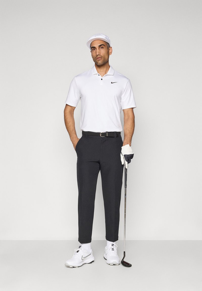 Man in white golf cap, white polo, black pants, and white golf shoes holding putter with gloved right hand, standing against plain background.
