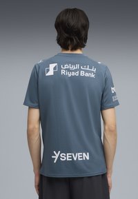 Camisa atlética gris con mangas cortas, textura suave y logotipos en blanco. Marcas visibles: Banco Riyad y SEVEN.