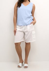 Haut sans manches bleu clair avec un décolleté en V, associé à un short cargo blanc avec des poches latérales. Des mules ouvertes blanches complètent la tenue.