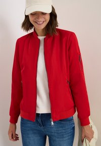 Femme portant une veste bomber rouge par-dessus une chemise blanche, un jean bleu et une casquette crème, souriant et tenant un sac de couleur claire dans sa main droite.