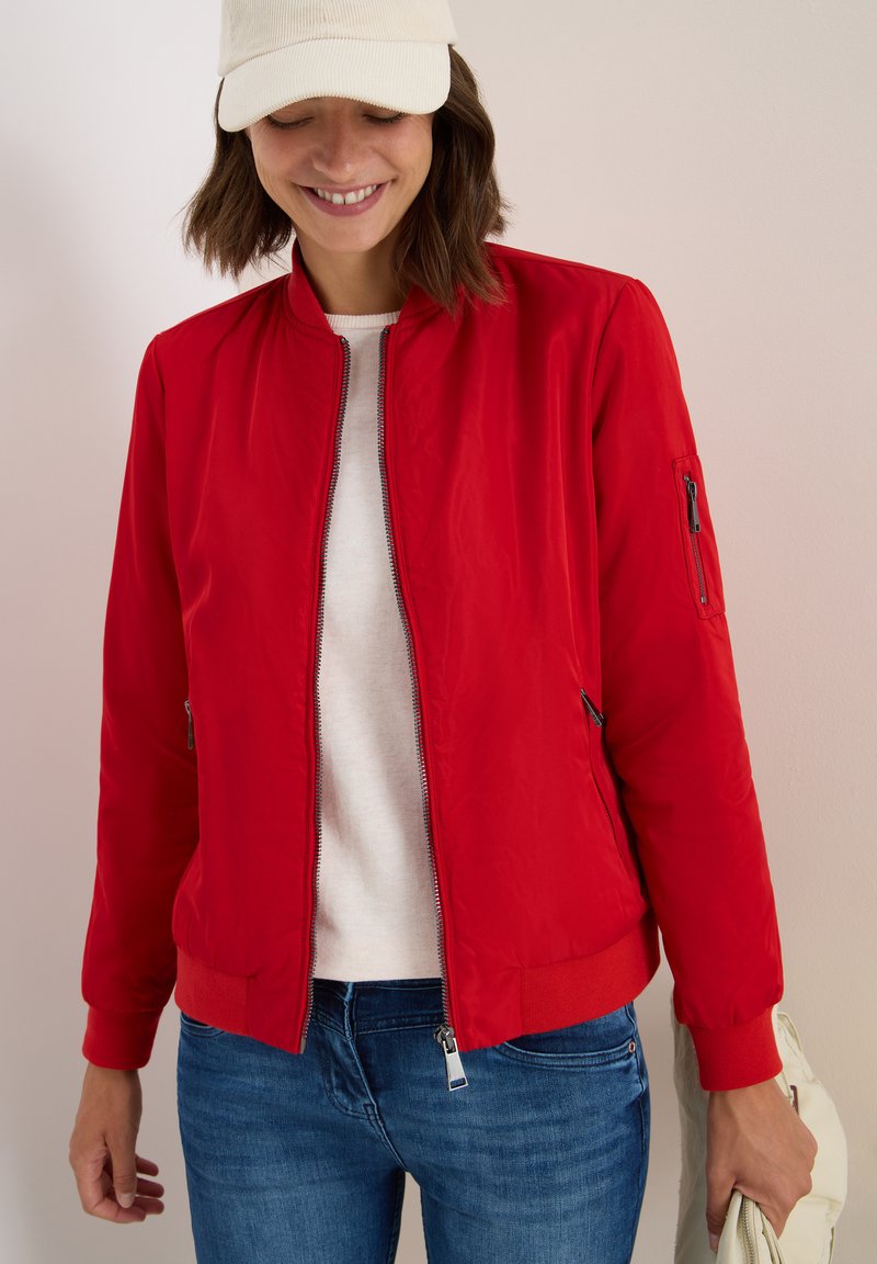 Femme portant une veste bomber rouge par-dessus une chemise blanche, un jean bleu et une casquette crème, souriant et tenant un sac de couleur claire dans sa main droite.
