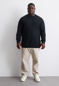 Schwarzer Pullover mit langen Ärmeln, beigefarbene Hose und weiße Sneakers. Der Pullover hat einen Rundhalsausschnitt; die Hose ist weit geschnitten und hat eine strukturierte Oberfläche.