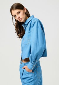 Klippt denimjacka i ljusblått, med knäppning och bröstficka. Tyget har en slät yta med subtila sömnadsdetaljer.