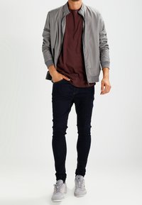 Grå bomberjacka över en marsala t-shirt, i kombination med mörka smala jeans och ljusgrå sneakers. Slät tyg och avslappnad design.