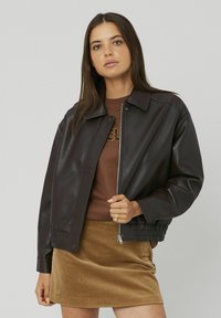 Veste bomber en cuir marron avec une fermeture éclair à l'avant, un col à pression et un ourlet élastique. Portée par-dessus une chemise marron et une jupe en velours côtelé beige.