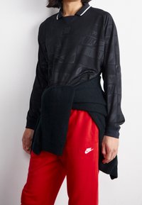Polo noir à manches longues avec un motif subtil, détails de col blanc, associé à un pantalon de survêtement rouge arborant un logo blanc, et un pull sombre noué à la taille.