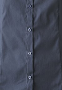 Camicia grigio scuro con una consistenza liscia, caratterizzata da una colonna di sei bottoni sul davanti, ognuno con un design circolare a contrasto.