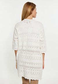 Bílý otvorený pletený cardigan s geometrickým vzorem, rukávy do lokte a vlnitý lem. Lehká a prodyšná textura.