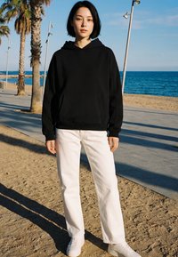 Jeune personne aux cheveux courts et noirs, portant un sweat à capuche noir, un pantalon blanc et des baskets blanches, debout sur une plage de sable près des palmiers et de la mer.