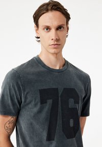 Czarny t-shirt z bawełny w ciemnoszarym kolorze z dużym czarnym nadrukiem liczby "76" na przodzie. Okrągły dekolt i krótkie rękawy. Miękka faktura.