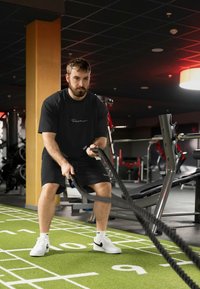 T-shirt et short d'entraînement noirs ; tenant des cordes épaisses noires sur un sol de gym vert texturé ; équipements de gym modernes en arrière-plan.
