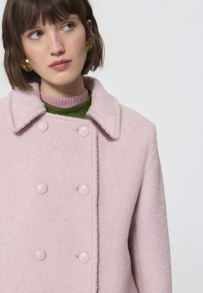 Cappotto di lana rosa chiaro con superficie testurizzata, dotato di colletto, design doppiopetto e bottoni rosa. Visibile sovrapposizione verde e viola.