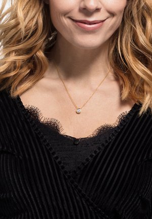 Collana d'oro con un ciondolo rotondo, indossata sopra un top nero a coste con rifiniture in pizzo lungo la scollatura. Elementi di design testurizzati ed eleganti.