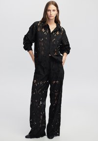 Completo in pizzo nero composto da una camicia a maniche lunghe, dal taglio ampio e con motivi floreali, abbinata a pantaloni larghi, che mettono in risalto una texture trasparente.