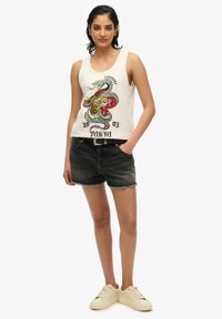 Beżowy top na ramiączkach z kolorową grafiką smoka i tygrysa, w połączeniu z czarnymi szortami dżinsowymi i beżowymi sneakersami. Prosty, codzienny design.